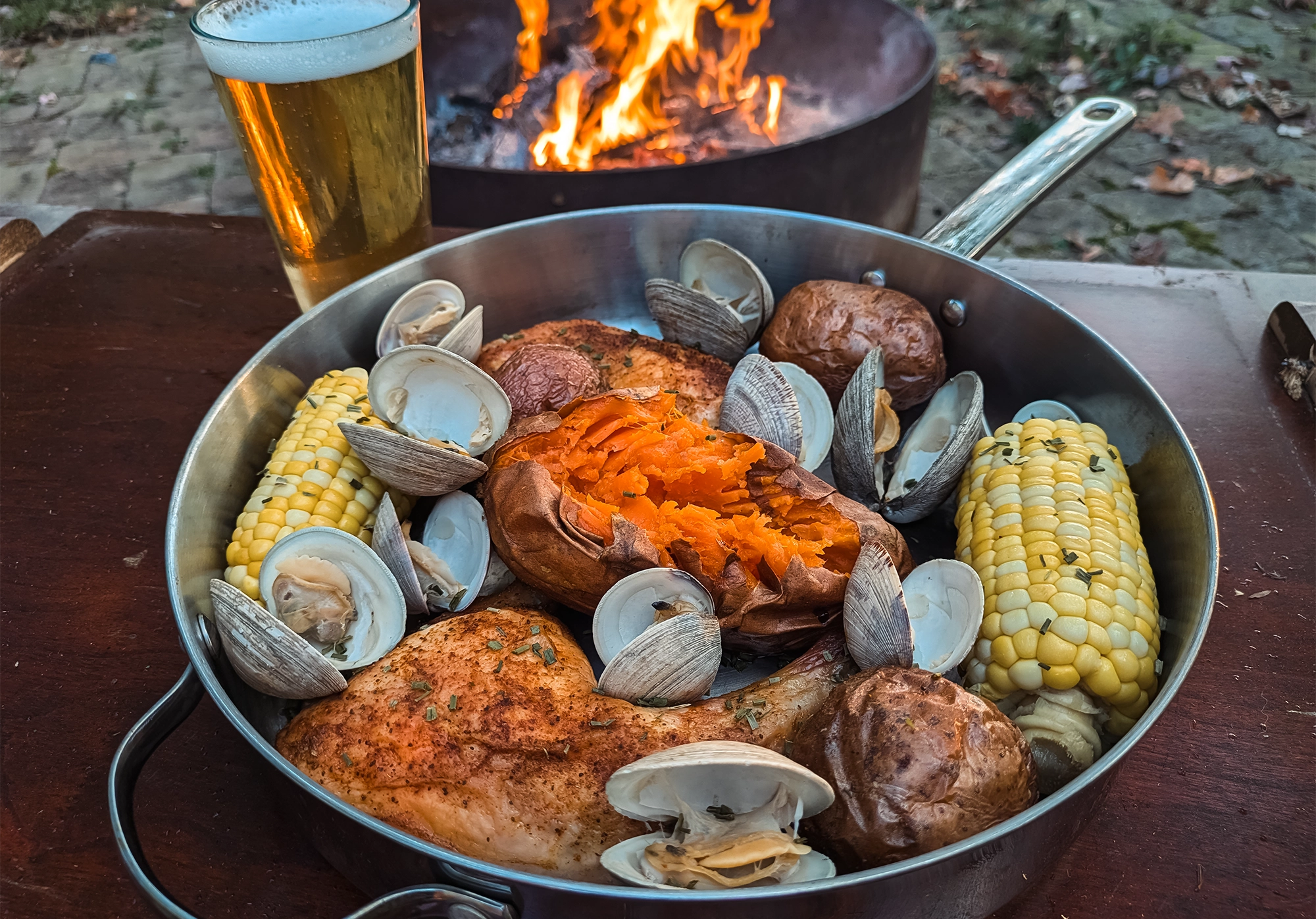 A pan of steamed clams, corn, chicken, and sweet potato beside a backyard fire pit.